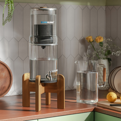 Clear water filter on a wooden stand with a glass pitcher filled with water on a kitchen counter.
